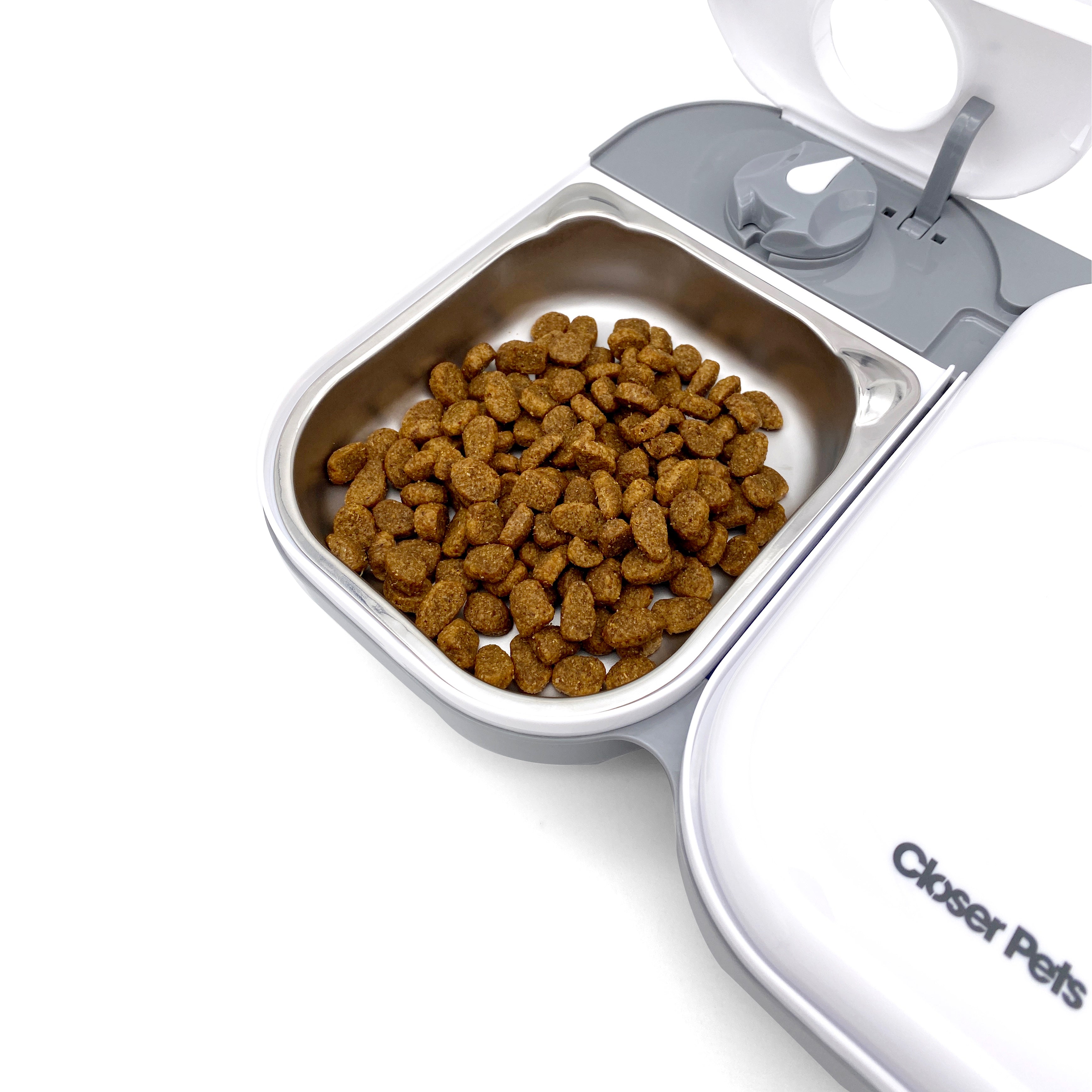 A grey and white two meal pet feeder with one bowl closed and another bowl filled with brown dry pet food in it on a white background
