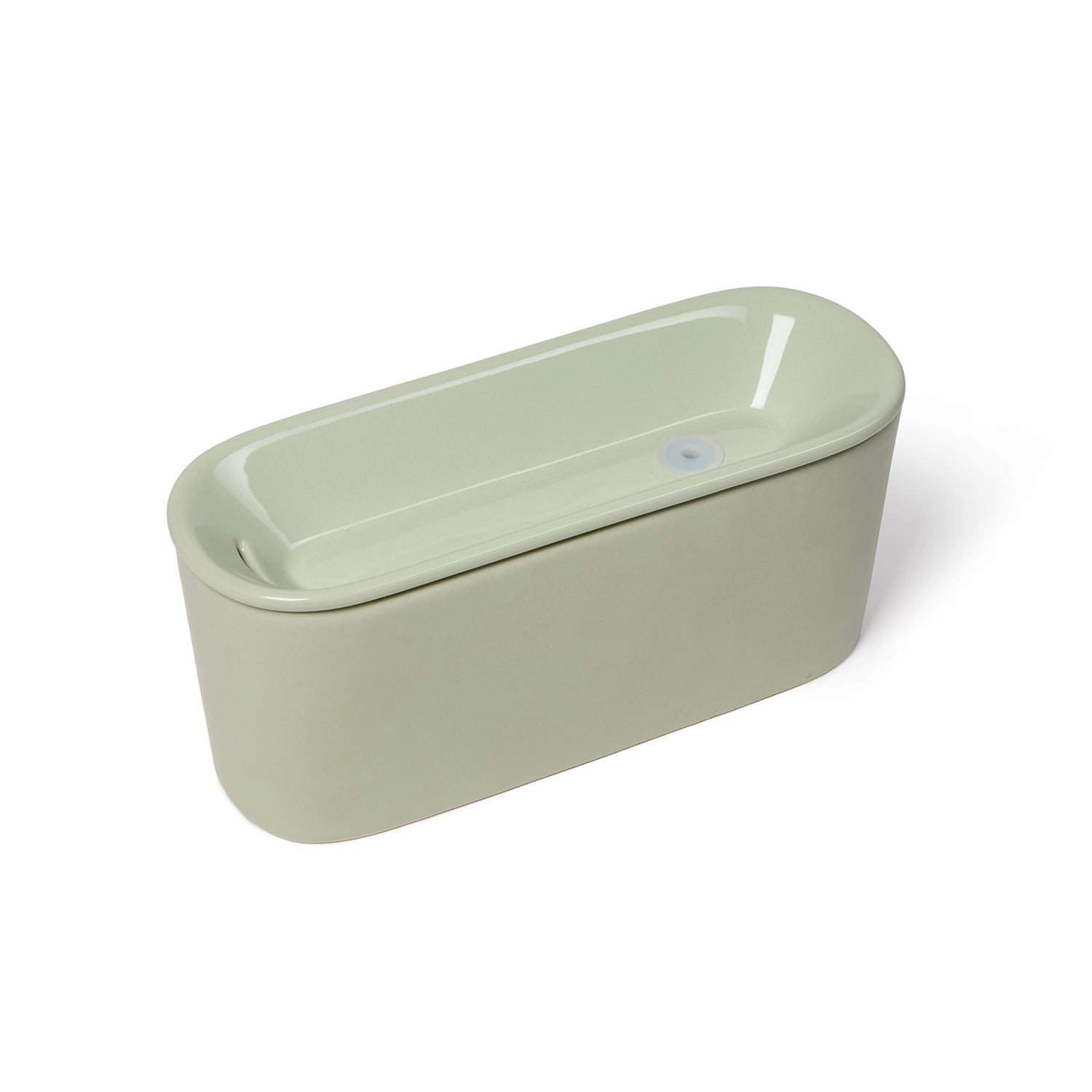 Photo of a sage green ceramic pet water fountain with a sleek, oval-shaped design. The fountain features a glossy finish with a lid that has an indented well where water flows. The minimalist design shows the compact, rectangular shape from an angled top view against a white background.