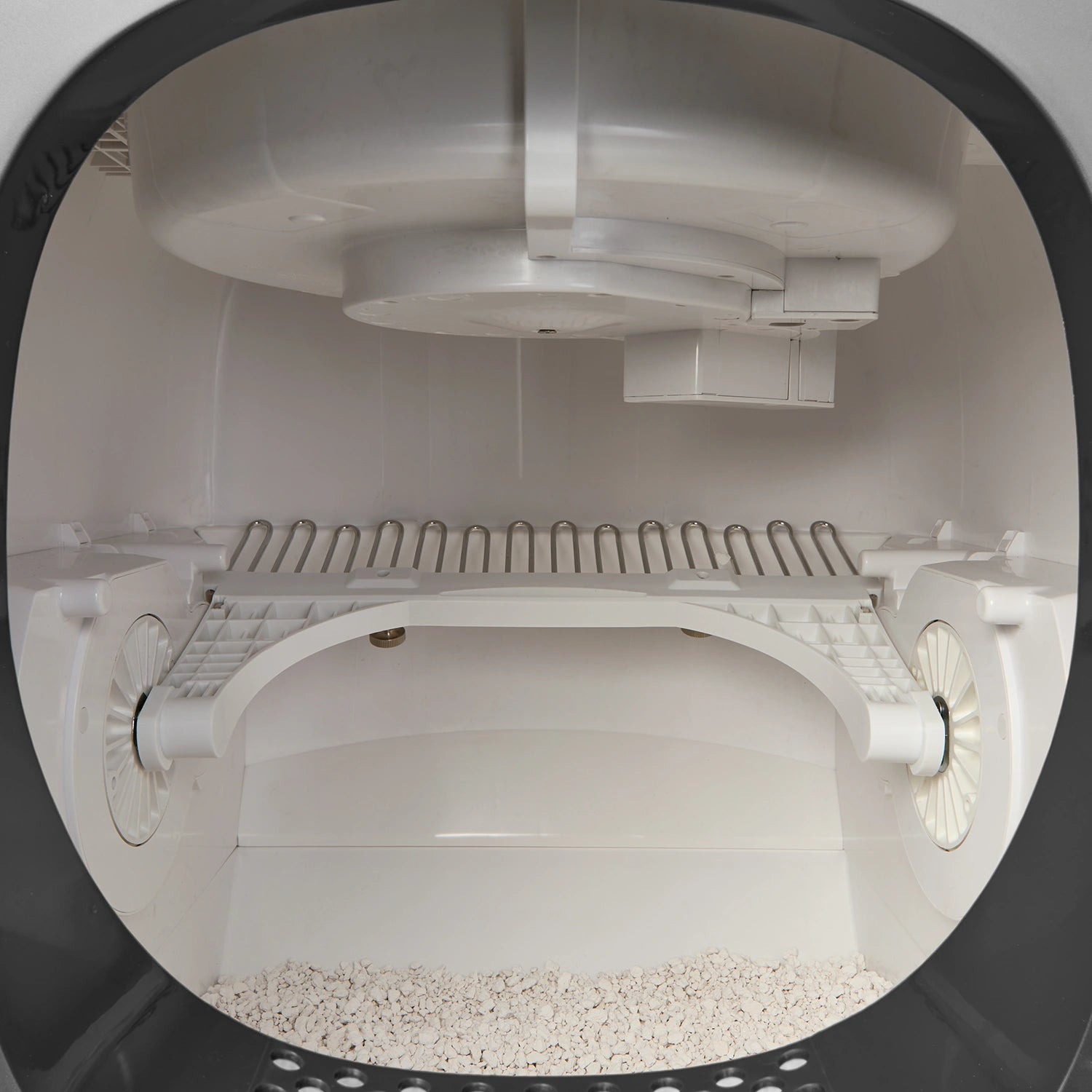 Interior view looking into the Cat Mate automatic cat litter box showing the self-cleaning mechanism with a white rotating cleaning rake mounted on cylindrical rollers. The rake features curved metal tines designed to sift through litter. Above is a white litter dispensing system with multiple compartments. Fresh clumping litter is visible at the bottom of the white chamber, viewed through the rounded entrance opening framed by a black rim.