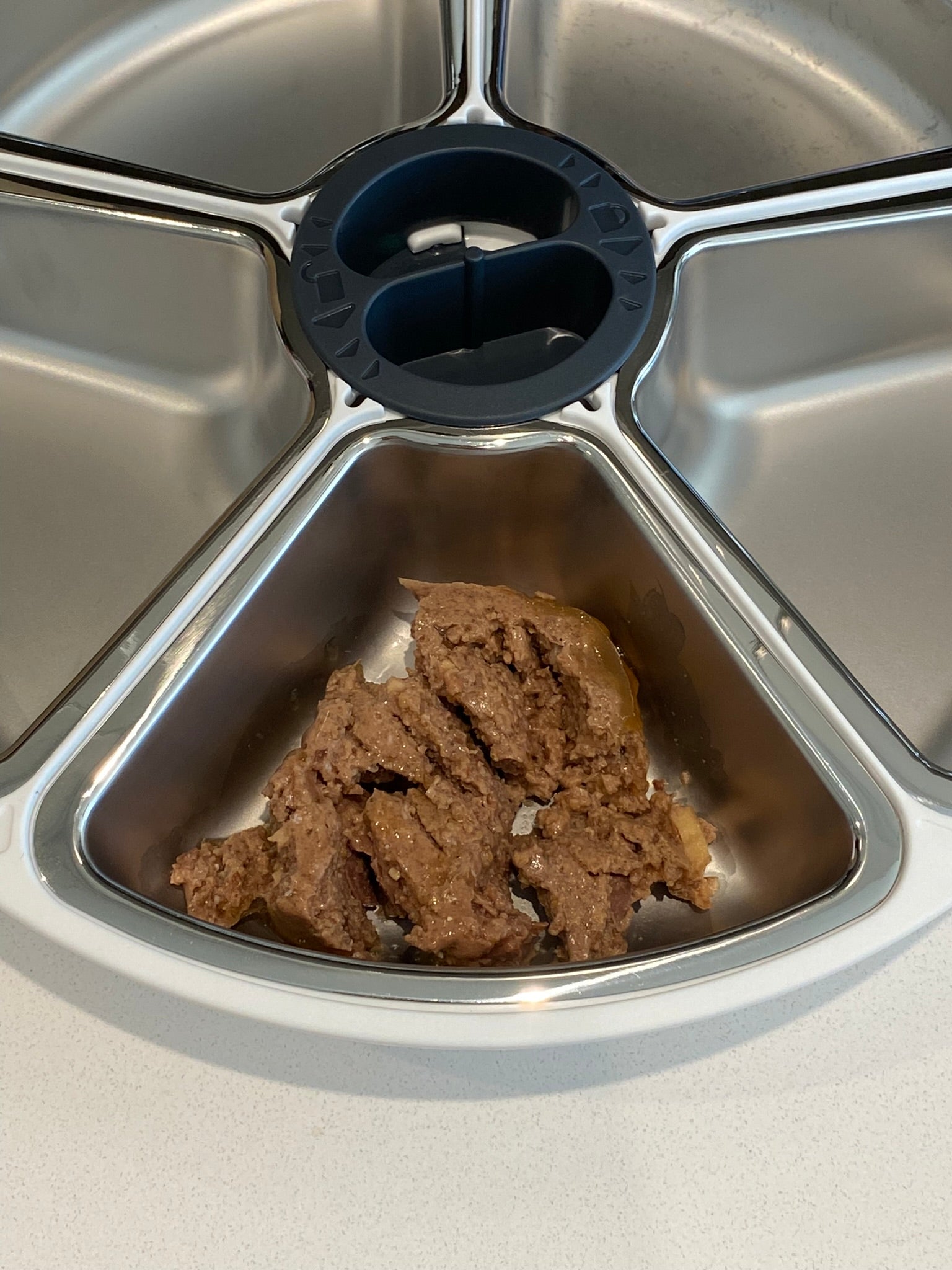 A portion of brown wet pet food in jelly placed in the nearest bowl insert within the ring of five stainless steel bowl inserts on a white background