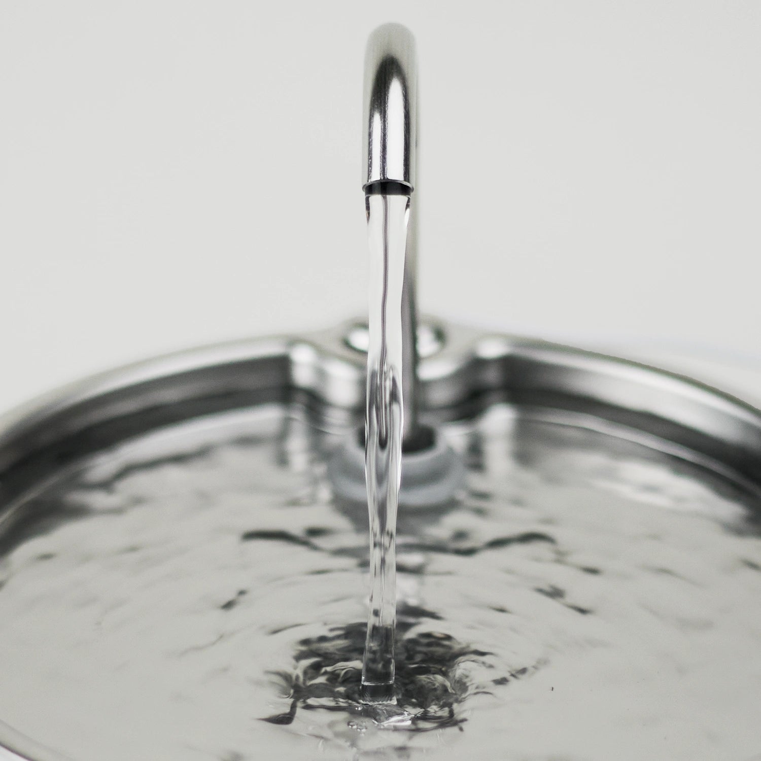 A close-up of a polished metal faucet spout dispensing a clear stream of water into the Cat Mate stainless steel fountain which is ideal for cats who prefer the flow of water from tall faucets spouts, shown against a light neutral background.