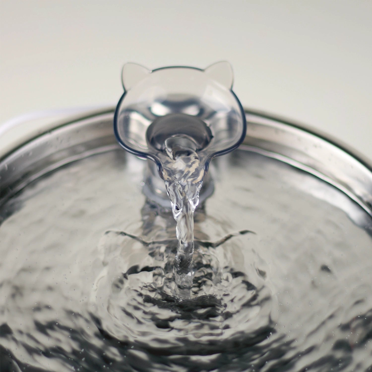A close-up of an acrylic cat-shaped faucet spout resting in the center of a stainless steel Cat Mate pet water drinking fountain, with water flowing into the bowl and ripples spreading across the surface.